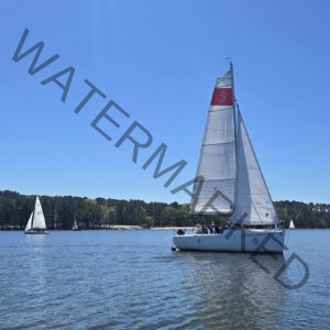 A white keelboat (a Beneteau First 22) sails across open lake water under full sail, its mainsail marked with a red panel and a seahorse-style logo. Two smaller sailboats and a low, wooded shoreline with a sandy bank sit in the background under a clear blue sky.