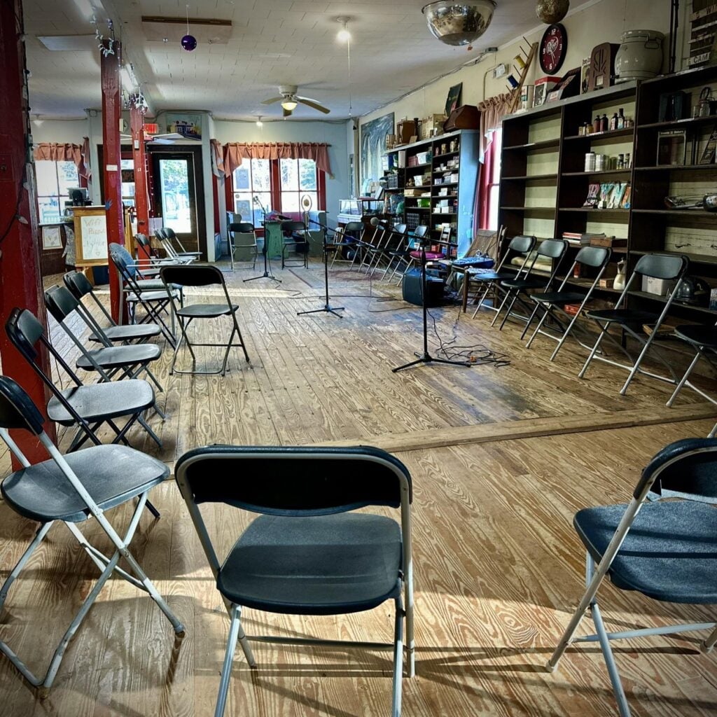 The inside of the Bynum General Store arranged for a performance: black and gray metal folding chairs form a wide ring on a worn wood-plank floor, surrounding several microphone stands and a small sound mixer. The walls hold tall wooden shelves filled with vintage store goods and bric-a-brac, with curtained windows, a ceiling fan, and a "Wifi Password" sign near the front door.