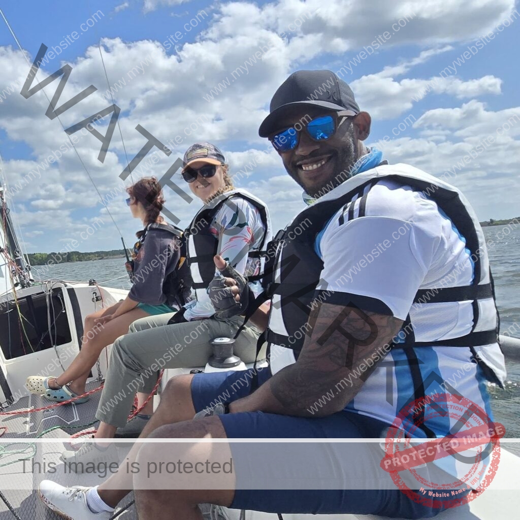 Three people wearing life jackets sit balanced along the side rail of a sailboat under a bright, partly cloudy sky, with open water and a distant tree line behind them. One holds a handheld radio; the group looks relaxed and at ease on the water.
