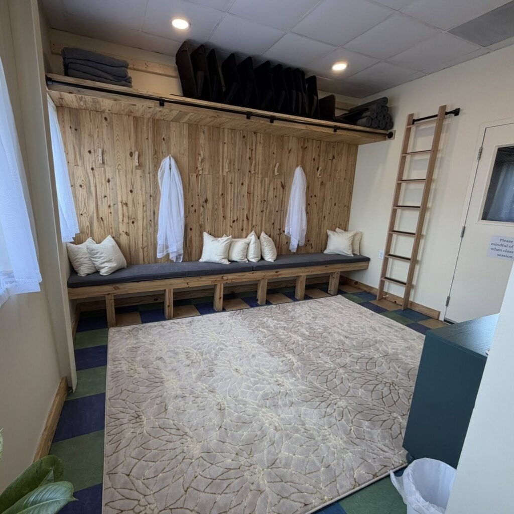 An interior lounge and prop-storage area inside Pittsboro Yoga. Against a natural pine-paneled wall sits a long wooden bench with a gray cushion and several cream throw pillows. Two sheer white robes hang from wall hooks, and a shelf running the length of the wall above holds folded blankets, rows of black yoga bolsters, and rolled mats. A tall wooden decorative ladder leans against the wall to the right, beside a door with a small window and a partly visible sign asking visitors to be mindful when class is in session. The floor is laid in a green-and-blue checkerboard pattern, mostly covered by a large cream rug with a subtle floral design. A leafy plant sits in the foreground corner.