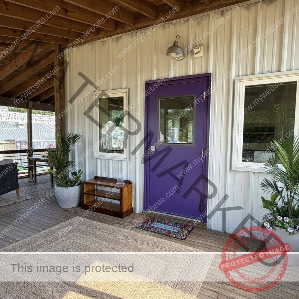The covered exterior entrance to Pittsboro Yoga at The Plant in Pittsboro. A bright purple door with a small window and a keypad lock sits in a cream metal-sided wall, flanked by white-framed windows and lit by a gooseneck barn light. A stained-glass-style "Welcome" mat lies on the wood-plank deck, with a woven area rug in front and potted palms and purple flowers on either side. The porch is shaded by an exposed wood-beam roof; to the left, a seating area with string lights and a view of other buildings across The Plant campus is visible.