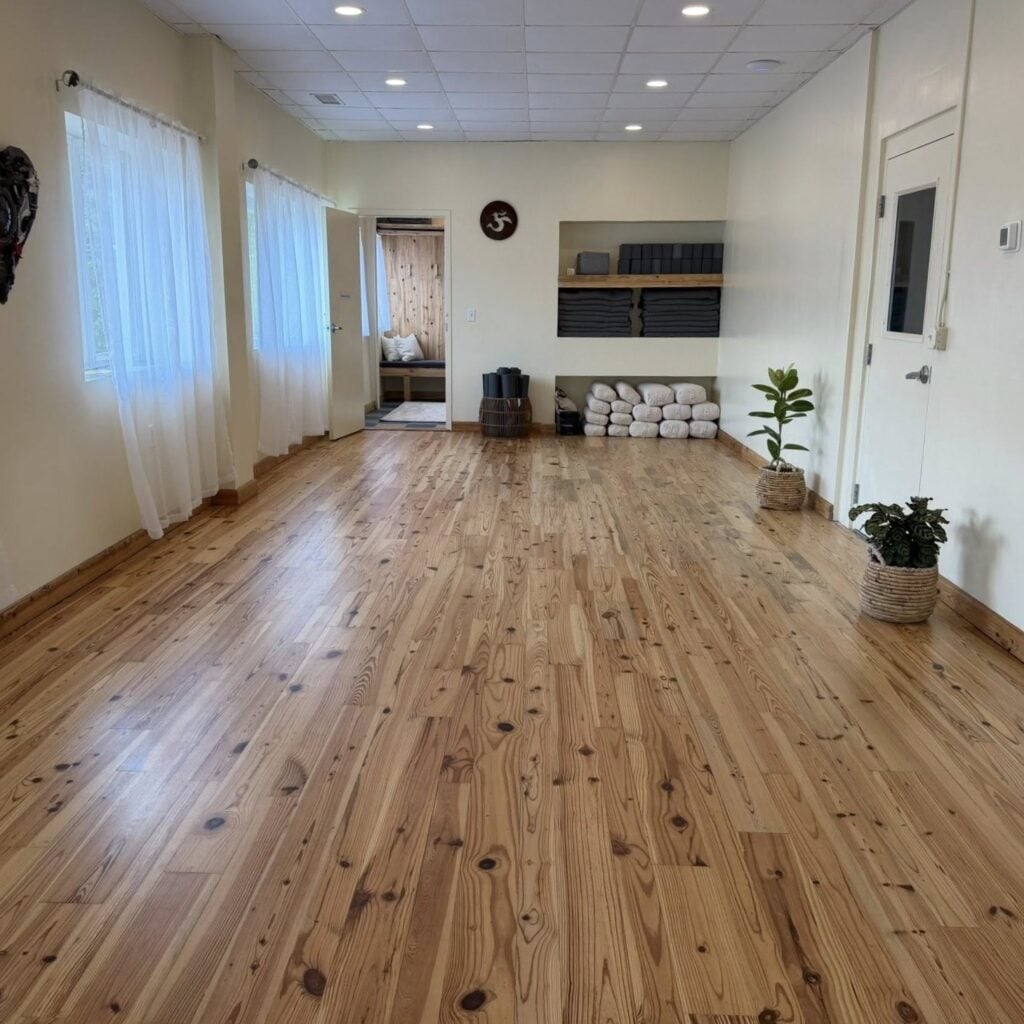The main practice room at Pittsboro Yoga, an open and airy studio with warm reclaimed-pine wood floors and cream walls. Tall windows along the left wall are dressed with sheer white curtains, and recessed lights run across a drop ceiling. A built-in wall niche on the right holds neatly folded gray blankets and stacked yoga blocks, with white bolsters and rolled mats arranged on the floor below and a couple of potted plants nearby. A round Om symbol hangs on the wall, and an open doorway at the back leads into the wood-paneled lounge area. The room is empty of people, showing the full practice space.