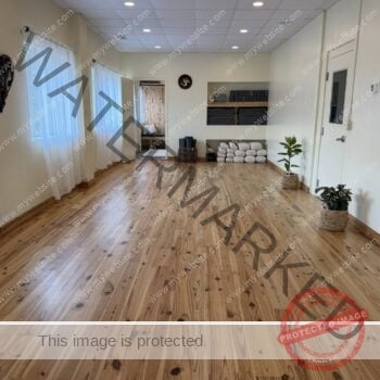 The main practice room at Pittsboro Yoga, an open and airy studio with warm reclaimed-pine wood floors and cream walls. Tall windows along the left wall are dressed with sheer white curtains, and recessed lights run across a drop ceiling. A built-in wall niche on the right holds neatly folded gray blankets and stacked yoga blocks, with white bolsters and rolled mats arranged on the floor below and a couple of potted plants nearby. A round Om symbol hangs on the wall, and an open doorway at the back leads into the wood-paneled lounge area. The room is empty of people, showing the full practice space.