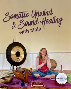 A promotional image for a Pittsboro Yoga class titled "Somatic Unwind & Sound Healing with Maia" in decorative purple script across the top. A person sits cross-legged on the floor, hands pressed together at the chest in a prayer position, eyes closed and smiling. They're surrounded by sound-healing instruments: a large bronze gong on a stand to the left, a wooden box drum, metal singing bowls, a crystal, small bottles, and other tools arranged on a purple mat in the foreground. The setting has pale yellow walls with white wainscoting and warm wood floors. The round Pittsboro Yoga logo appears in the lower-right corner.