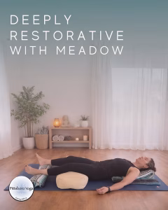 A promotional image for a Pittsboro Yoga restorative class, titled "Deeply Restorative with Meadow" in large text across the top. A person lies on their back on a blue yoga mat in a relaxed restorative pose, eyes closed, with the backs of the knees draped over a cream bolster and the head and shoulders resting on folded blankets and patterned pillows. The setting is a calm, light-filled studio with warm wood floors, sheer white curtains over tall windows, a potted plant, and a low wood console holding a small Buddha figure, rolled towels, a candle, and greenery. The round Pittsboro Yoga logo appears in the lower-left corner.