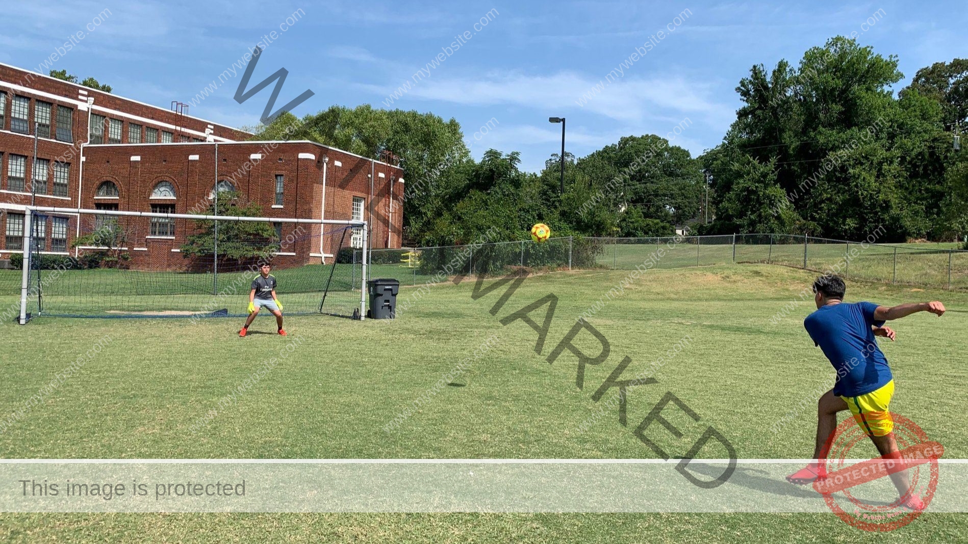 Two people play soccer on an open grassy field on a sunny day. In the foreground at right, one player in a blue shirt and yellow shorts follows through on a kick, sending the ball airborne toward the goal. At left, a goalkeeper in a dark jersey and yellow gloves crouches ready between the posts of a portable goal. A large historic red-brick building with arched windows stands behind a chain-link fence, framed by mature green trees and a blue sky.