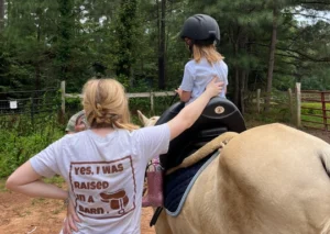A woman in a light t-shirt reading "Yes, I was raised in a barn" rests a hand on the back of a young child who sits in the saddle of a tan horse, wearing a riding helmet. They face a wooded paddock with a wooden fence and a red metal gate in the background.