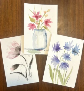 Three watercolor paintings on textured paper arranged on a dark wood surface, each signed "R. Brown." The top card shows pink wildflowers in a pale blue mason jar; the lower-left card features a single loose pink and black bloom with ink-brushed leaves; and the right card depicts a cluster of blue and purple cornflowers on green stems. All are painted in a soft, loose, wet watercolor style.