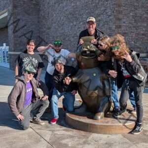Seven people pose and mug for the camera, clustered around a large bronze statue of a smiling cartoon pig seated on a round base. They wear casual clothes, hoodies, hats, and sunglasses, several striking exaggerated or rock-and-roll poses. A stone-faced building with vending machines is behind them on a sunny day.